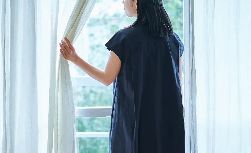 Woman, who is a nosy neighbour, standing by a window, looking outside through parted curtains.