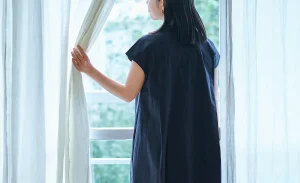 Woman, who is a nosy neighbour, standing by a window, looking outside through parted curtains.