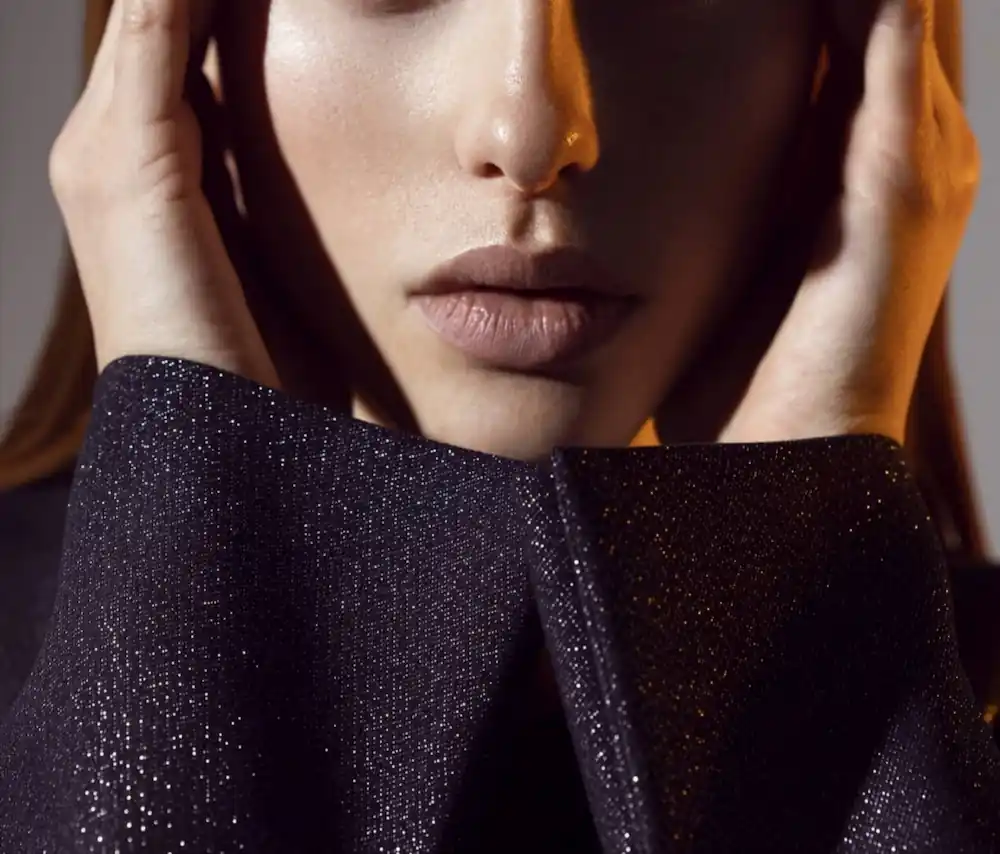 Close-up portrait of a sophisticated older woman wearing a shimmering black top, with her hands gently framing her face.